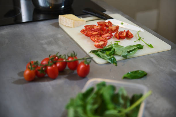 A kitchen countertop featuring freshly cut tomatoes, leafy greens, and cheese on a cutting board. Modern kitchen preparation scene highlighting fresh ingredients.