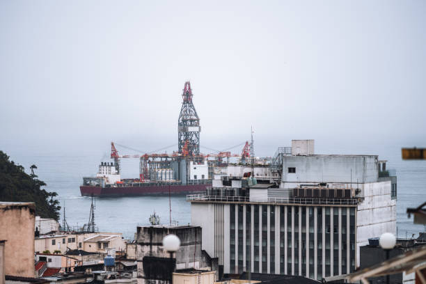 Offshore oil drilling platform is moving or anchored near a city coast, visible among urban buildings. The industrial rig stands out against the overcast sky, highlighting maritime and urban contrast