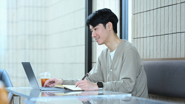 istock Smiling young man sitting at glass table in cafe, working on his laptop and writes in notebook 2164136974
