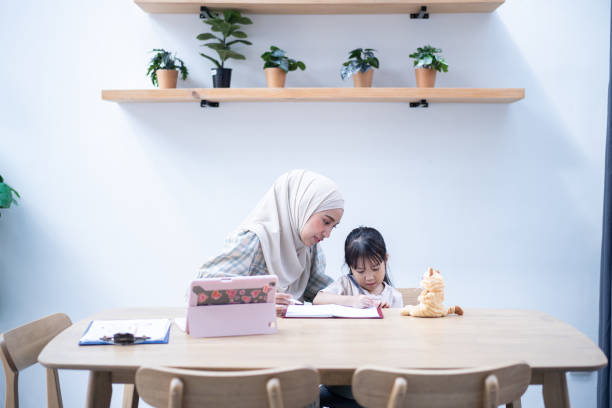 istock Mother working and  helping her daughter with homework at home while sitting at a wooden table with educational materials and a laptop 2164141925
