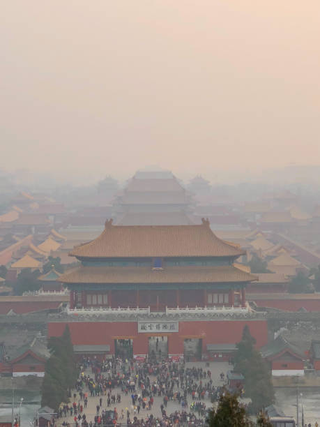 A view of the Forbidden City in Beijing, China, enveloped in heavy smog. The iconic imperial palace complex, with its traditional Chinese architecture, appears muted and distant through the thick haze. The bustling crowd at the entrance adds a dynamic element to the otherwise subdued and atmospheric scene, highlighting the enduring significance and historical importance of this UNESCO World Heritage Site despite environmental challenges.