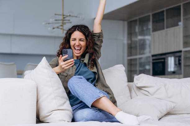 istock Joyful woman screams while looking at her smartphone with her hand raised, has won a gift or read the good news. 2164163477