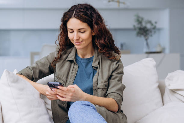 istock A woman sits relaxed on a sofa, deeply engrossed in her smartphone, epitomizing casual leisure in a serene home setting. 2164163763