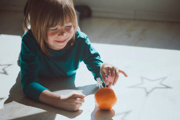 Cute portrait of a little boy playing an astronaut. He uses an orange as a planet or a Moon.  Hand-painted American flag. Landing on the Moon. Star-shaped shadows on the floor.