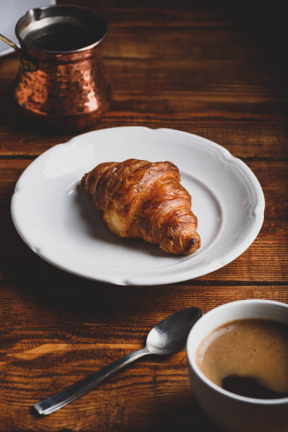 Stock photo showing elevated view of freshly baked breakfast pastry of all butter croissant on white plate besides a plate of biscotti, a metal teaspoon and a blue and white striped, china espresso cup and saucer full of black coffee on a grey muslin.