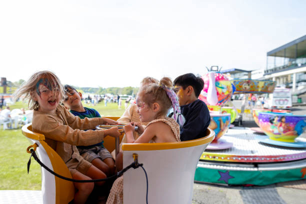 Group of children on a teacup fairground ride at an Easter fair in the North East of England. They are having fun, putting their arms up. Some of the children have their faces painted. One of the boys is looking at the camera while laughing.
