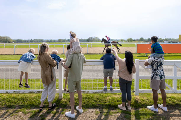 Rear view wide shot of two families visiting an Easter fair at a racecourse in the North East of England. They are standing side by side watching the horse race together, cheering it on. Some of the children are sitting on their father's shoulders.