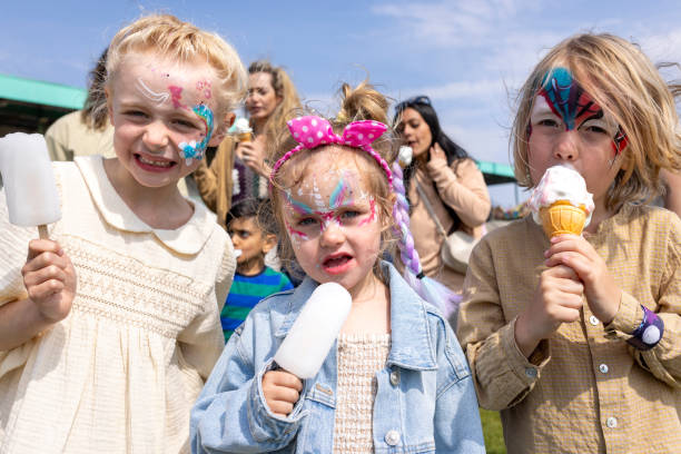 Children siblings standing looking at the camera together while visiting an Easter fair in the North East of England. They are eating ice creams and frozen lollies, enjoying the sweet treats. The children have their faces painted.