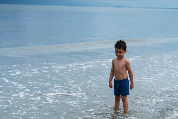 A lone child in a vibrant red swimsuit sits on the sandy beach, contemplating the crashing waves. Serenity surrounds