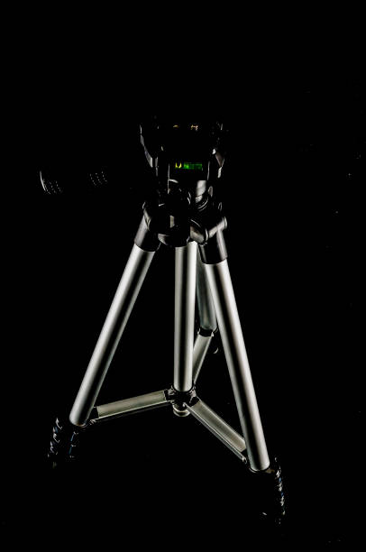 A tripod is standing on a black background. The tripod is silver and has a green lens