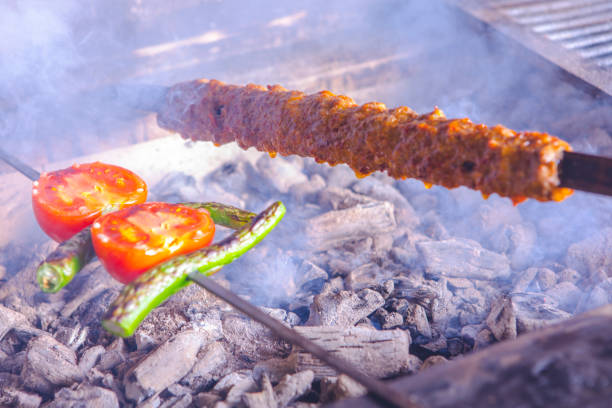 This photo shows a delicious shish kebab grilling over hot coals. The skewer contains red meat and green peppers, along with tomatoes. The smell of the meat and vegetables wafts through the air as the skewer rotates over the hot coals. The photo shows the preparation of a delicious dish in an appetizing way.