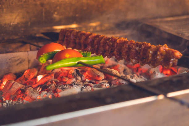 This photo shows a delicious shish kebab grilling over hot coals. The skewer contains red meat and green peppers, along with tomatoes. The smell of the meat and vegetables wafts through the air as the skewer rotates over the hot coals. The photo shows the preparation of a delicious dish in an appetizing way.
