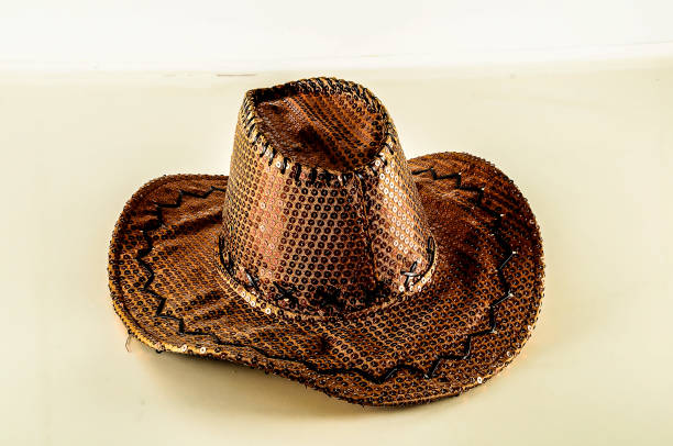 A brown cowboy hat with a black band sits on a white surface. The hat is made of straw and has a rustic, old-fashioned look