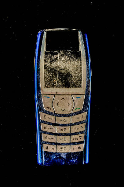 A cell phone with a blue case is shown on a black background. The phone is old and has a cracked screen. Scene is somewhat melancholic, as the phone appears to be discarded and no longer in use