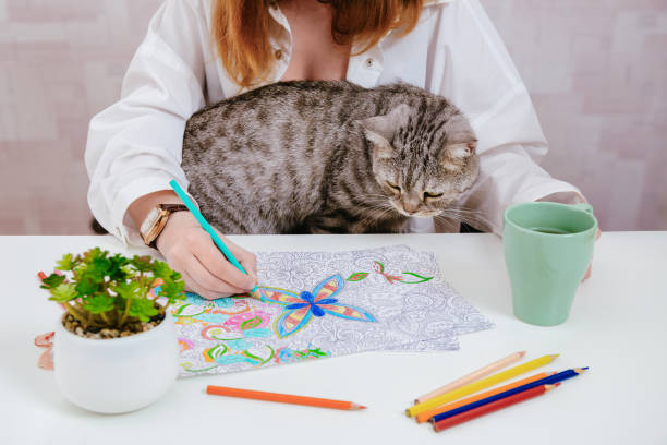 A woman colors a mandala or anti stress coloring pages while a gray cat sits on her lap.