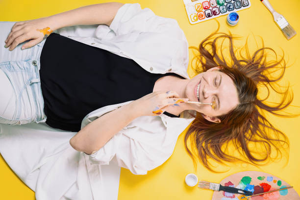 A smiling artist lies on a yellow background, holding a paintbrush. Paints, brushes, and palette are scattered around