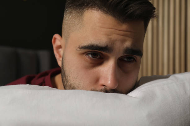 Serious man on blue background holding head on pillow and looking at camera, having trouble sleeping concept