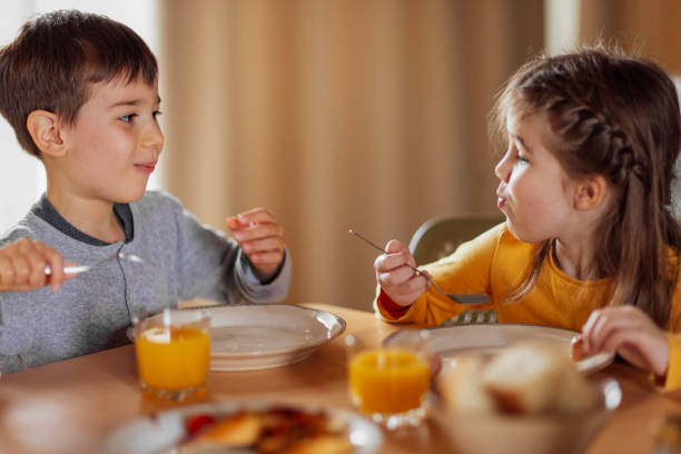 Two small children are sitting at dining table and having breakfast. Cute kids smile and look at camera. Lovely brother and sister in casual clothes are eating in kitchen at home. Happy childhood.