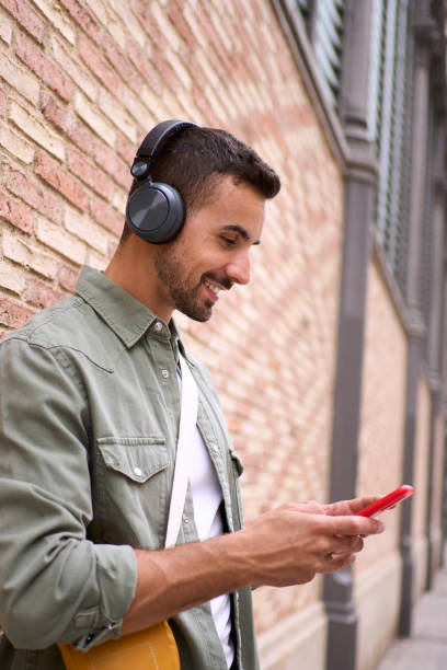 istock Attractive young Caucasian man with wireless headphones using cell phone leaning against brick wall. 2164265859