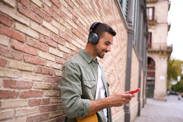 istock Attractive young Caucasian man with wireless headphones using cell phone leaning against brick wall. 2164271085