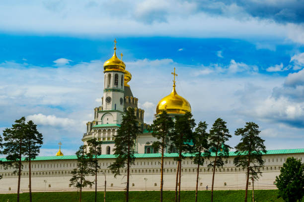 The golden domes of New Jerusalem Monastery rise above its white walls, set against a blue sky with trees in the foreground.