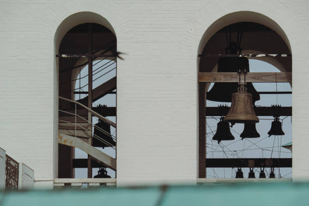 A close-up view of large bells hanging inside the arches of a white tower. The structure's interior details, including the wooden beams and supporting cables, are visible through the arches.