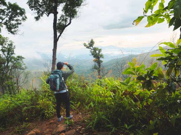 istock Hiker with backpack stands overlooking lush green valley shrouded in mist and clouds. Backpack has clear plastic water bottle in the side pocket. 2164301044