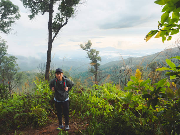 istock Hiker with backpack stands overlooking lush green valley shrouded in mist and clouds. Backpack has clear plastic water bottle in the side pocket. 2164301301