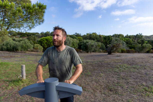 A man exercises on a stepper machine at an outdoor gym, surrounded by lush greenery. The image captures a moment of focused physical activity in a natural, serene environment.
