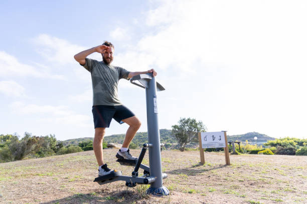 A man wipes sweat from his forehead after exercising on a stepper machine at an outdoor gym. The bright, open setting emphasizes a healthy and active lifestyle in nature.