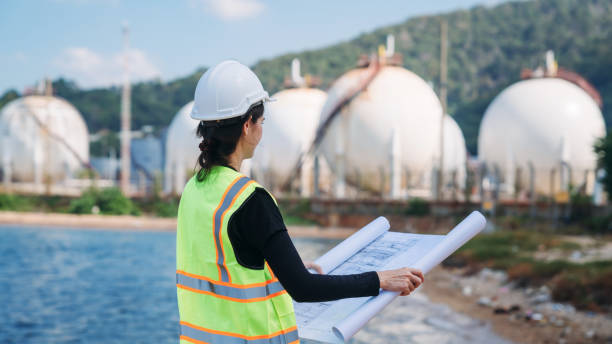 istock Assured engineer wearing a safety helmet and high-visibility vest holds blueprints, with industrial tanks in the background. 2164317401