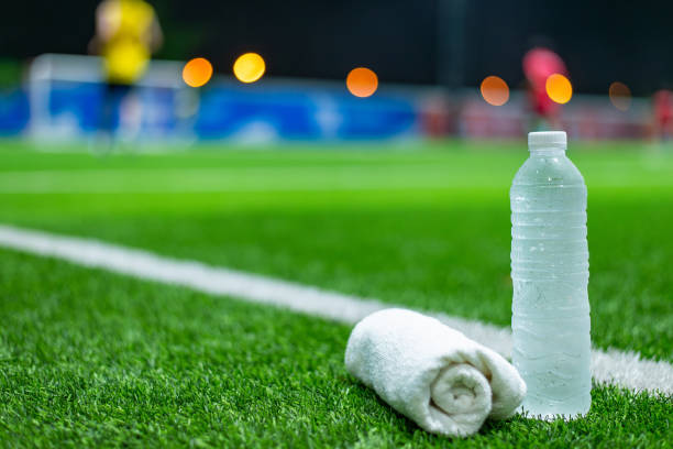 Close-up of a water bottle and towel on a soccer field, with blurred players in the background during night practice.