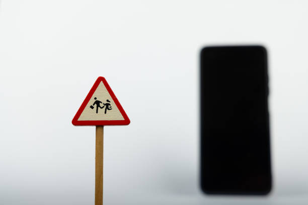 A wooden warning sign for children stands out in focus, while a smartphone appears blurred in the background, creating a simple white setting for emphasis