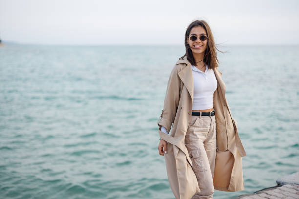 Portrait of happy young woman in long autumn coat walking on the sea dock