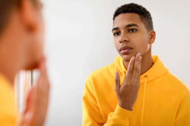 Black teen guy checking skin in mirror, looking thoughtful