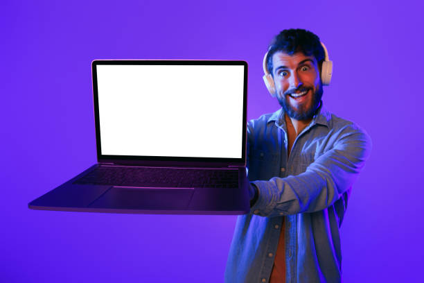 istock Smiling Guy In Headphones Shows Laptop With Mockup Screen, Studio 2164328324