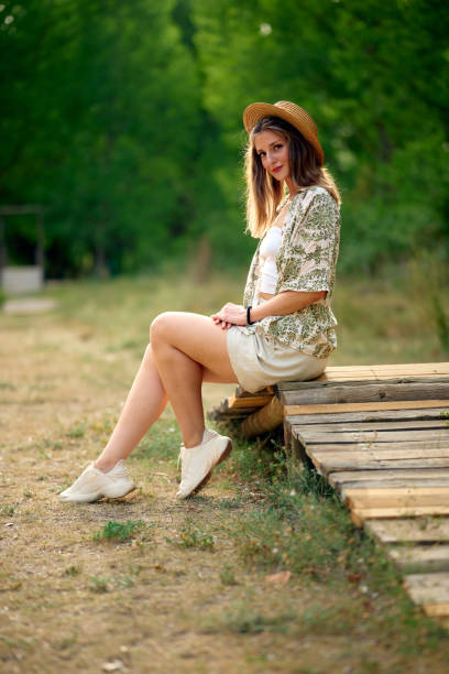 A young woman gracefully poses in a vibrant green forest, wearing a straw hat. The sunlight filters through the trees, blending effortlessly with the natural surroundings.