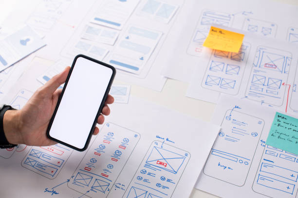 istock UX/UI designer working on wireframes for a website and mobile app prototype, surrounded by sketches of user-flow and design tools, in concept of website and mobile application design concept. 2164339339
