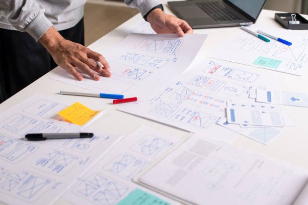 istock UX/UI designer working on wireframes for a website and mobile app prototype, surrounded by sketches of user-flow and design tools, in concept of website and mobile application design concept. 2164339358