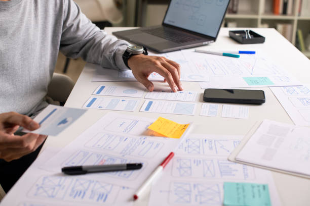 istock UX/UI designer working on wireframes for a website and mobile app prototype, surrounded by sketches of user-flow and design tools, in concept of website and mobile application design concept. 2164339363