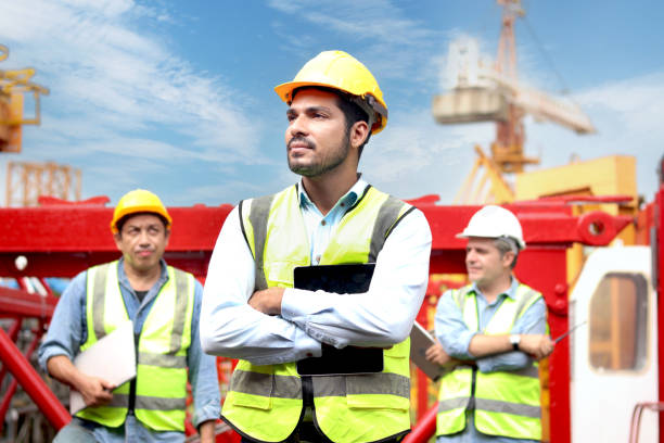 istock Portrait handsome engineer worker with safety vest and helmet holding tablet, standing with arms crossed with coworker as background at construction building site. young architect works at workplace. 2164352607