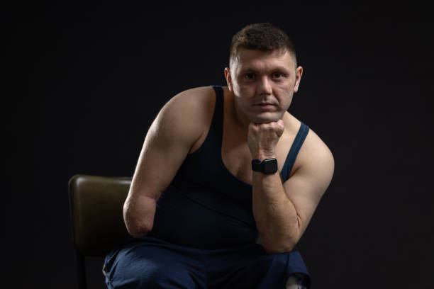A three-quarter length shot of a young male adult athlete sitting on a chair with his head balanced on his hand in a photographic studio. He is a quadruple amputee who developed Meningitis as a baby, looking into the camera with a positive expression on his face wearing navy blue shorts and a vest.