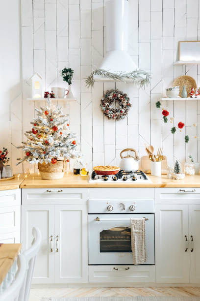 White scandinavian kitchen with Christmas decorations. A snow-covered Christmas tree with garland lights and red and silver Christmas toys on the kitchen countertop. Christmas interior
