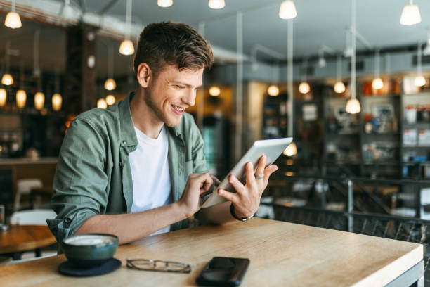 Young Man Reading and Using Digital Tablet in Bookstore Young man sitting in modern bookstore ,library, reading and using his digital tablet cafe tablet stock pictures, royalty-free photos & images