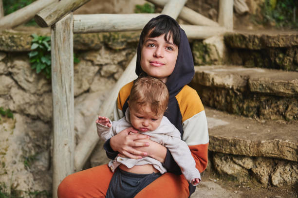 A loving scene of an older brother holding his baby sibling while sitting on stone steps in a rustic outdoor setting. The older boy wears a hoodie, creating a cozy family moment.