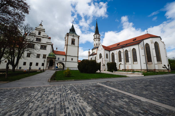 A historic church with a tall spire and an adjoining building stand beside a cobblestone square under a partly cloudy sky. The architecture features traditional elements, and the scene is framed by trees and well-maintained greenery.