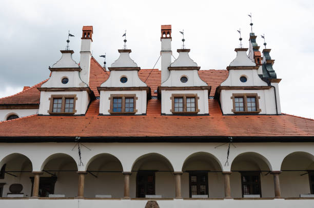 A historic building featuring a red gabled roof with multiple dormer windows and intricate weather vanes. The structure includes an arched colonnade below, showcasing classic architectural details under a partly cloudy sky.
