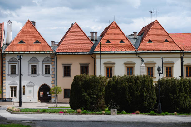 A row of traditional townhouses featuring red gabled roofs and ornate facades, located in a quaint town square. The scene is framed by manicured hedges and a cobblestone path, set under a partly cloudy sky.