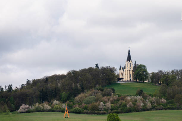 A picturesque church with a tall spire is situated atop a hill, surrounded by dense trees and greenery. The serene landscape under a cloudy sky creates a peaceful and idyllic scene.