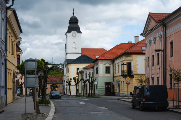 A Baroque-style church with an ornate facade and clock tower stands in a town square. The church is surrounded by other historic buildings and parked cars, under a cloudy sky, highlighting the town's rich architectural heritage.